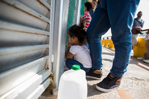 A three-year-old from Honduras peers through a fence at the U.S. - Mexico border while her family waits to apply for asylum. Photo by Jesse Costa for WBUR