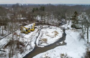 Traphole Brook, flowing away from the camera and across the dam at the right side of the picture. The demolition team dug the smaller channel that runs to the left and through the berm beyond the construction equipment. The team will divert the brook along this channel before they remove the dam.