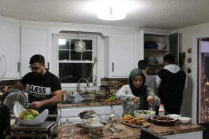 Mashan Manahe, left, transfers a meat dish from the stove while his wife, Zainab, makes a salad and their two sons, Omar and Nasir, get plates out of the cabinet. The Manahe family enjoys a variety of specialty food during Ramadan, including dates, sambousas, and sweets.