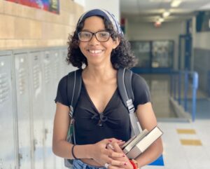 A photo of a female high school student with short, dark wavy hair and glasses in the hallway of her school with lockers in the background. The student is wearing a backpack and holding books in her arm.