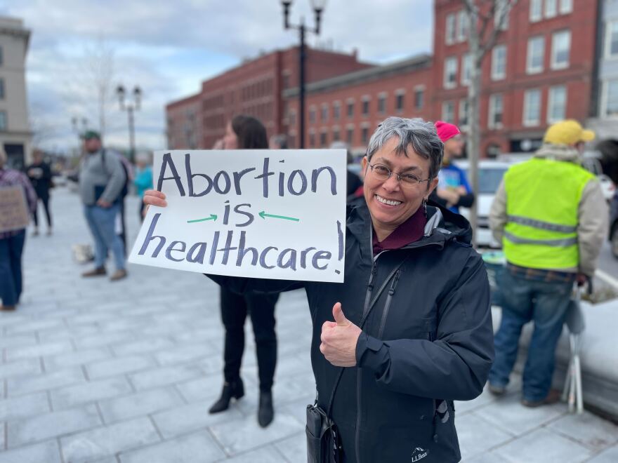 An older woman wearing eyeglasses smiles and stands on a sidewalk, giving a thumbs up while holding a protest sign that reads, "Abortion is healthcare!"