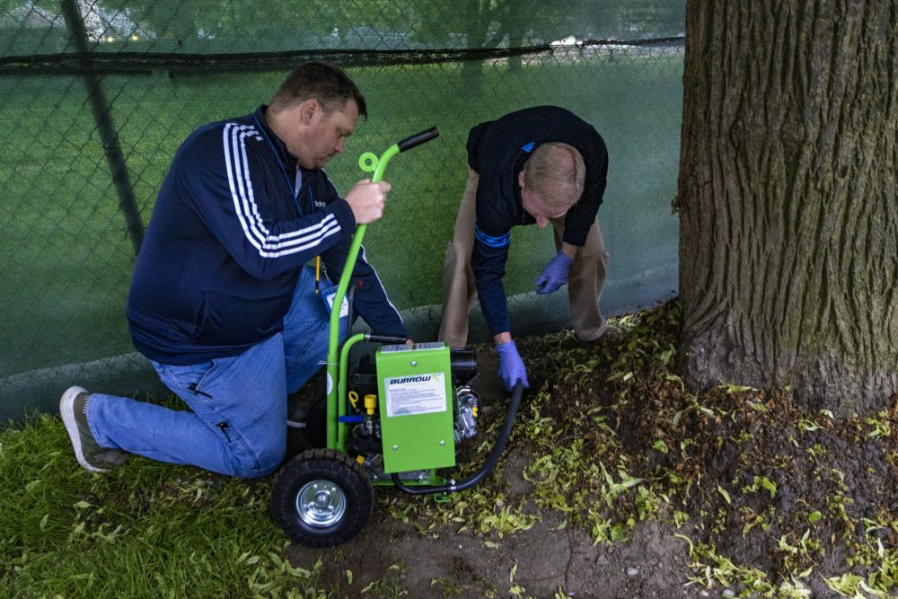 A man wearing a navy Adidas track jacket kneels next to another man and a tree as they use a machine called BurrowRx to pump carbon monoxide into a rat burrow on Boston Common. of Boston Environmental Sanitation Division, place a hose from the Burrow RX into a hole of a rat burrow and begin pumping carbon monoxide into it. 