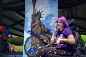 A young bespectacled woman with purple hair down to her shoulders, sits in a motorized wheelchair in front of a vertical banner that's a painting of the Statue of Liberty in a wheelchair.