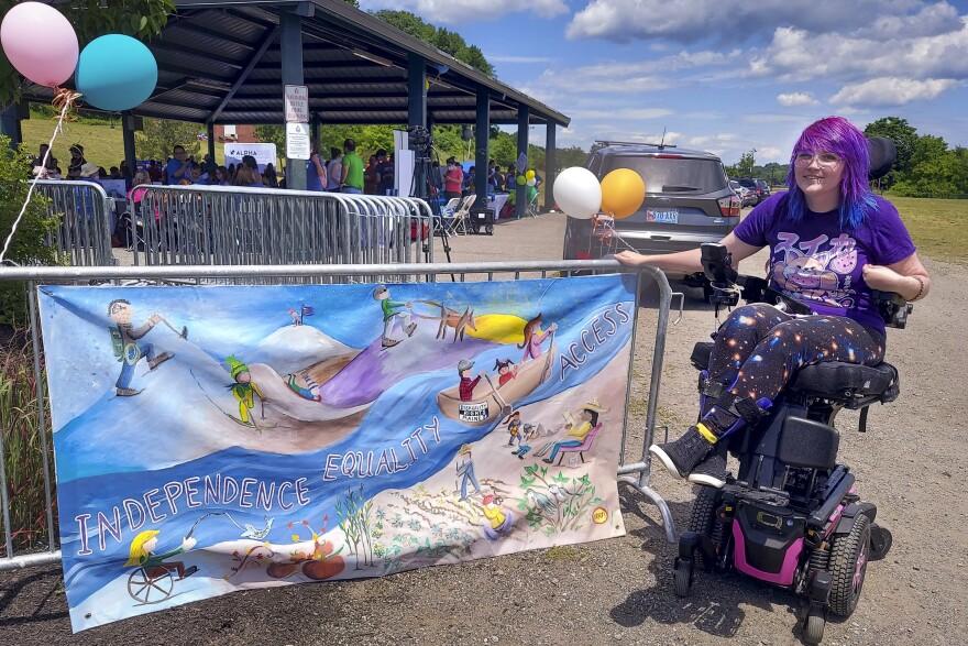 A young bespectacled woman, with purple straight hair just past her shoulders, wears a colorful outfit and sits in a motorized wheelchair next to a banner that states "Independence Equality." She is sitting outside at an event and there are clouds in the sky.