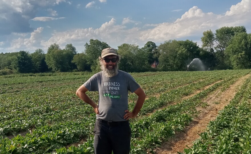 Ryan Voiland, owner of Red Fire Farm in Granby and Montague, Mass., wears a tan cap, sunglasses and stands in front of a field of crops. He has a beard and poses with his hands on his hips.