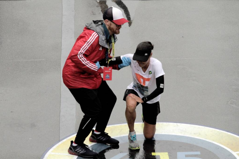 A member of the marathon's medical team helps a runner stand up from a kneel just across the finish line at the 2023 Boston Marathon. The runner is kneeling on the 4.15 insignia, which commemorates the Boston Marathon bombing a decade ago.