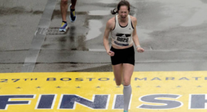 Leah Gaeta, a woman wearing bib number 8828, screams in triumph as she crosses the yellow finish line of the 2023 Boston Marathon.