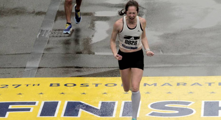 Leah Gaeta, a woman wearing bib number 8828, screams in triumph as she crosses the yellow finish line of the 2023 Boston Marathon.