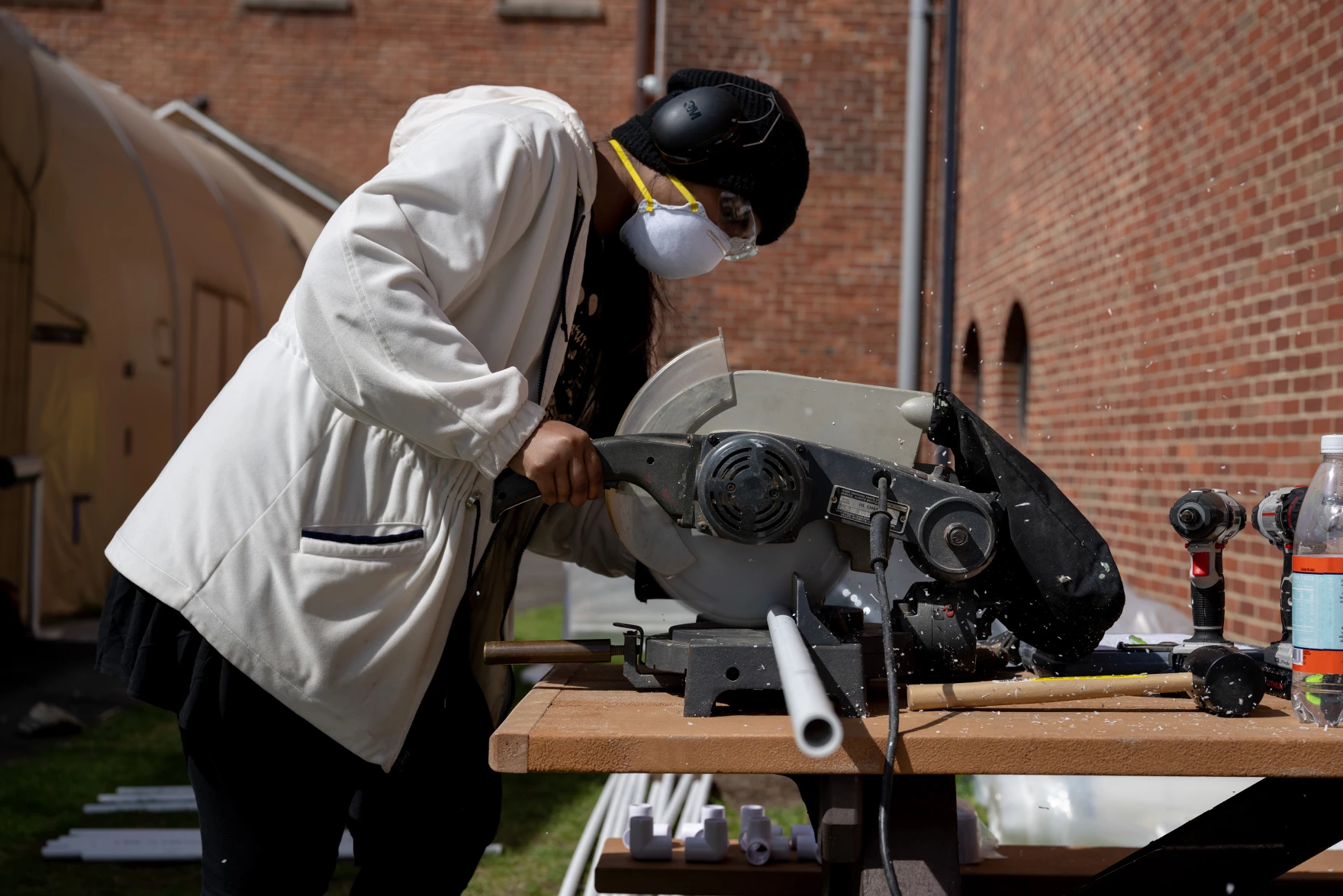 Rebha Raviraj, wearing a white hoodie, black head covering, white face mask and safety googles, uses a power saw to cut PVC pipe behind the the Maritime Aquarium in Norwalk, Conn. The researchers will set up greenhouses at eight salt marshes across Connecticut.