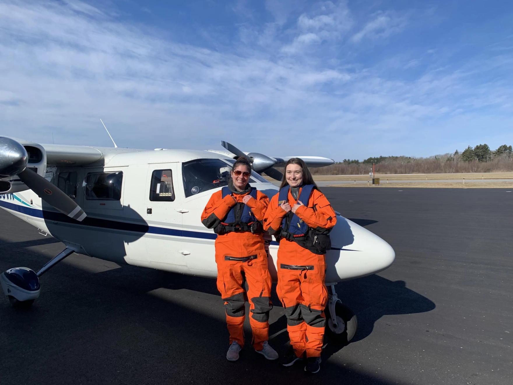 Orla O'Brien and Katherine McKenna, wearing orange jumpsuits and standing in front of a small plane, are both scientists at the New England Aquarium and prepare for takeoff from the New Bedford Regional Airport on March 10, 2023. The two will fly over the Nantucket Shoals as part of the aquarium's aerial survey program, which documents North Atlantic right whales, dolphins, sharks and other marine mammals. 