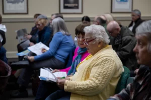 Nashua residents Pat Lyons, an older woman with short white hair and wearing a yellow cardigan sweater, and Irene Oliveira, wearing a pink sweater with a blue-ish scarf, are seated at a city meeting and look at informational sheets on April 5, 2023, during a meeting about community power at City Hall in Nashua, N.H.