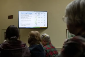 A TV screen shows "Electricity Choices" with text explaining various options. It's shown to Nashua residents attending a meeting on April 5, 2023, about community power at City Hall in Nashua, N.H.