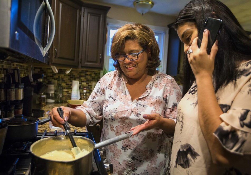 As Irma Flores stirs mik in a pan, her daughter Gabriela Portillo-Perez speaks with her grandmother Isabel on the phone in El Salvador. Photo by Jesse Costa for WBUR.
