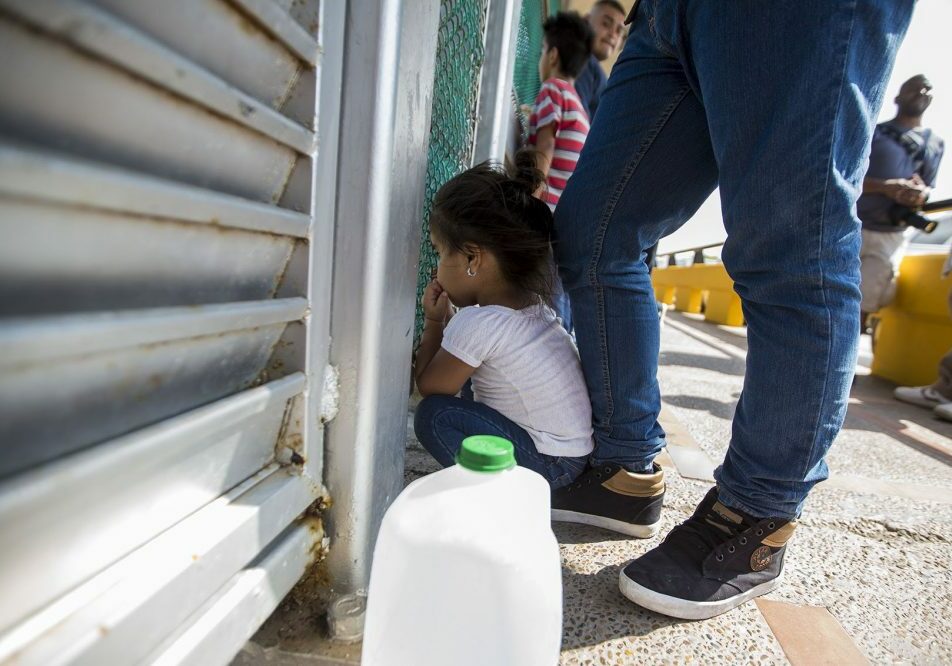 A 3-year-old from Honduras peers through a fence at the U.S.-Mexico border while her family waits to apply for asylum. Photo by Jesse Costa for WBUR